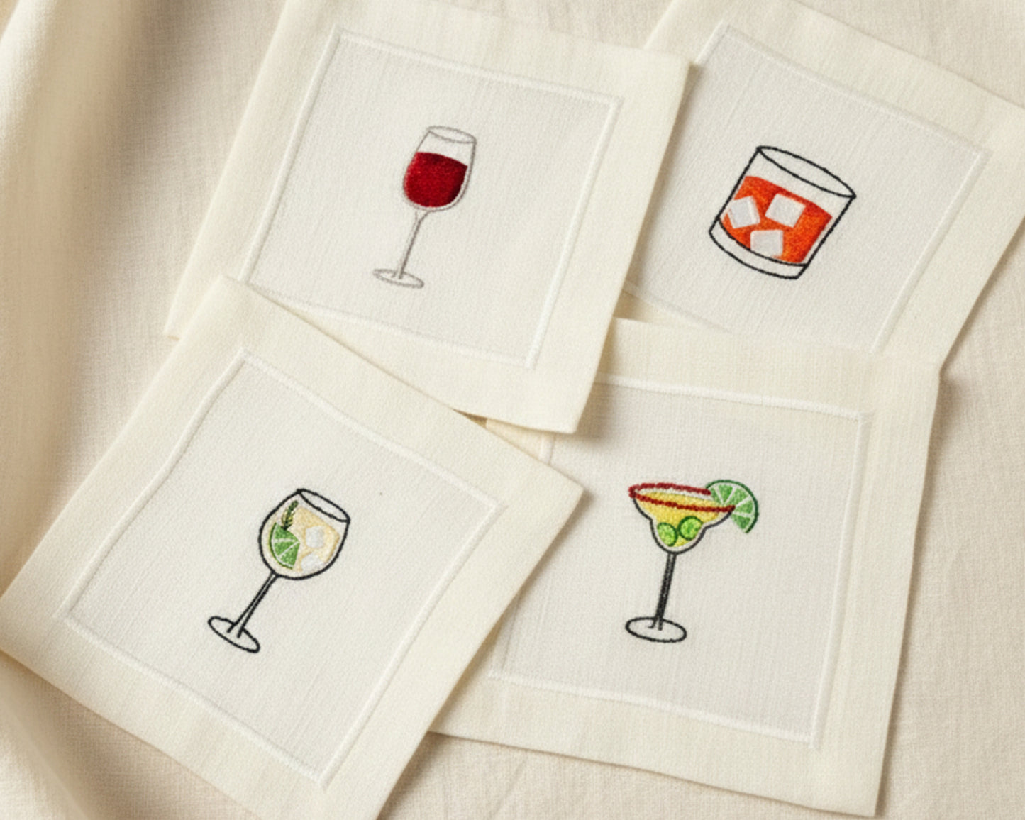 Close-up of four cotton napkins with colorful embroidery including an Aperol Spritz, Blue Margarita, Martini, and Espresso Martini.