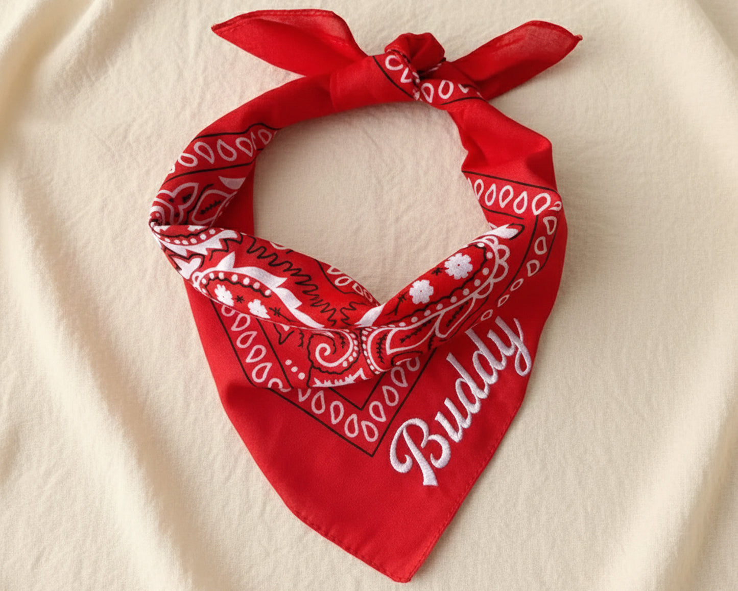 A stack of four folded paisley bandanas in burgundy, cream, black, and light blue, each featuring a unique name in chain stitch script.