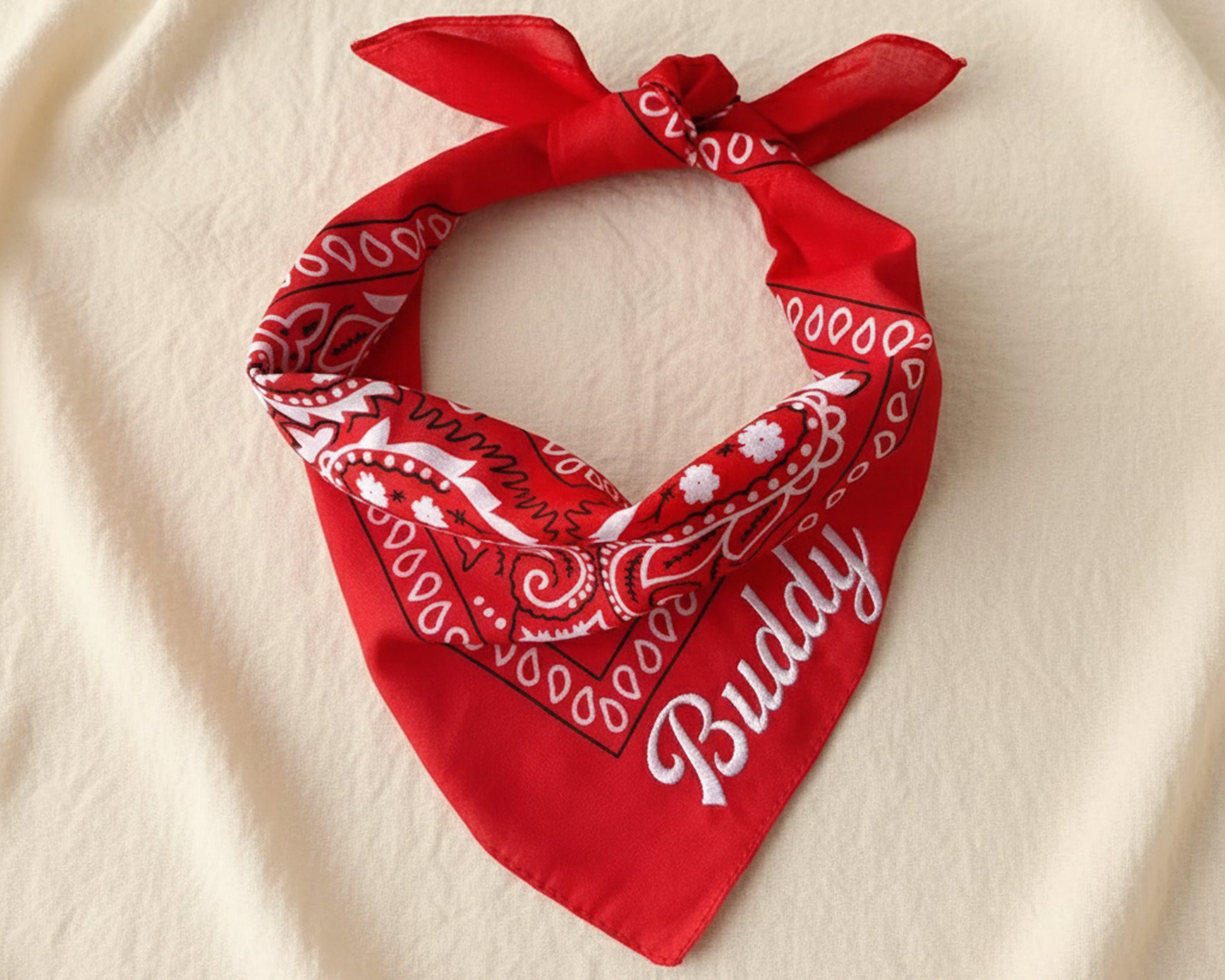 A stack of four folded paisley bandanas in burgundy, cream, black, and light blue, each featuring a unique name in chain stitch script.