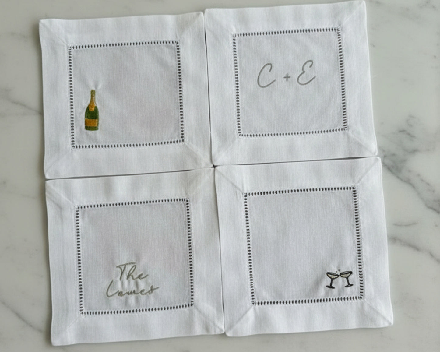 Flat lay of four personalized white linen coasters featuring champagne bottle, initials, name, and martini icons