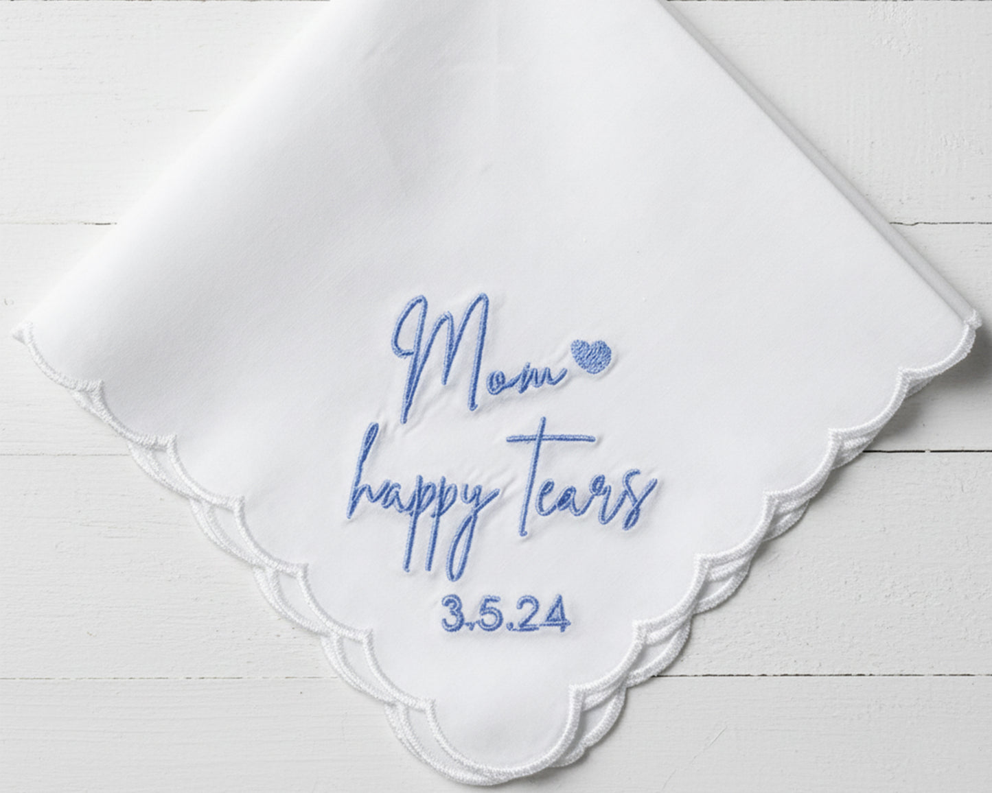 A close-up of a premium white cotton handkerchief with a wave-stitched scalloped edge and custom blue embroidery for "Mom happy tears".