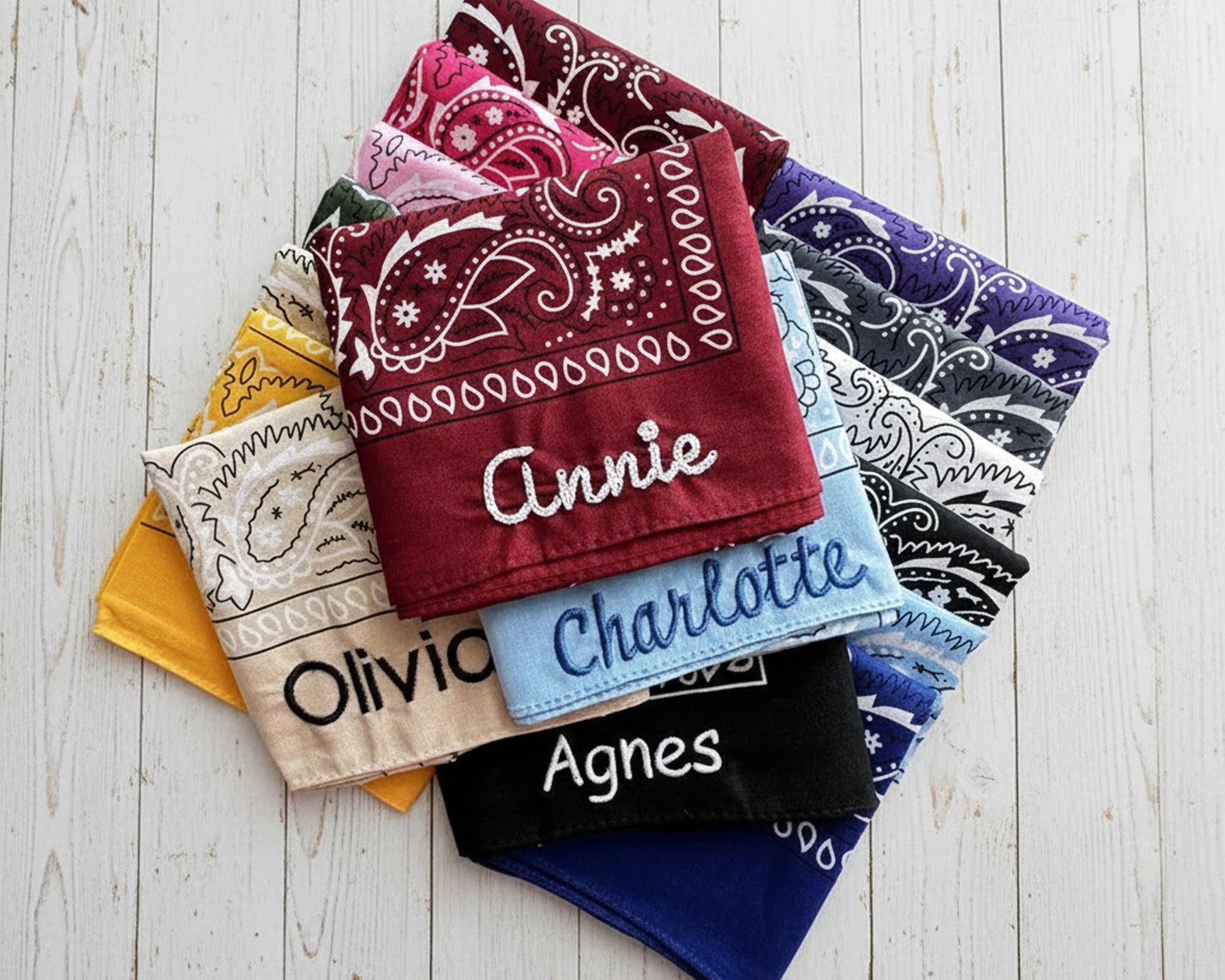 A stack of colorful paisley bandanas in red, blue, black, and pink featuring personalized names like Annie, Charlotte, and Olivia in chain stitch embroidery