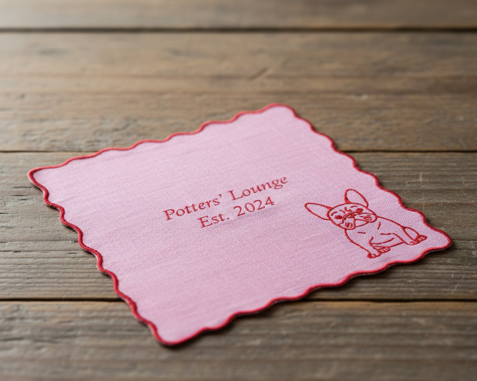 A lilac pink linen cocktail napkin with a red scalloped edge, personalized with "Potters' Lounge Est. 2024" and a French Bulldog.