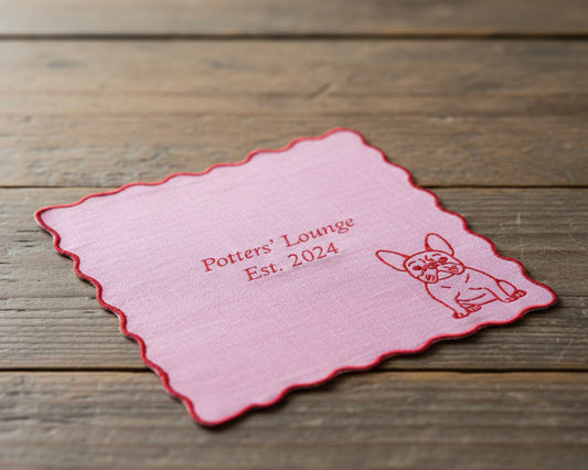 A lilac pink linen cocktail napkin with a red scalloped edge, personalized with "Potters' Lounge Est. 2024" and a French Bulldog.