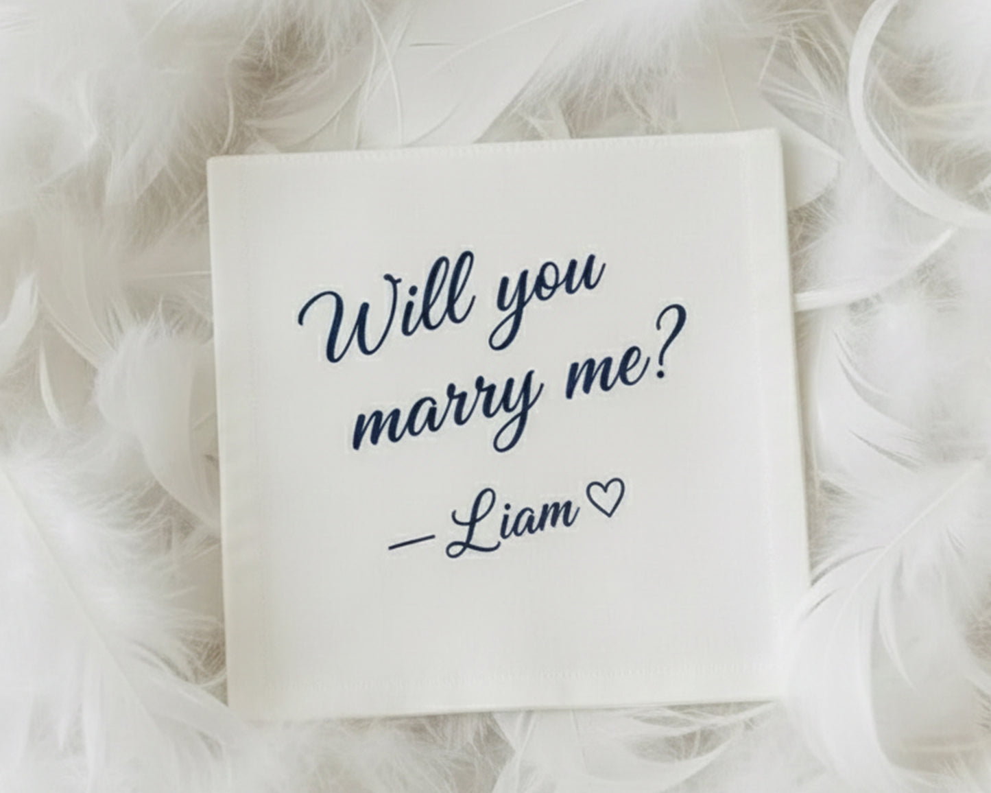 A personalized white proposal handkerchief with navy vinyl text asking "Will you marry me? — Liam ❤" surrounded by soft white feathers.