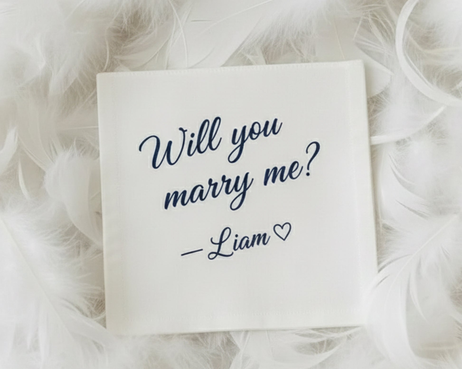 A personalized white proposal handkerchief with navy vinyl text asking "Will you marry me? — Liam ❤" surrounded by soft white feathers.