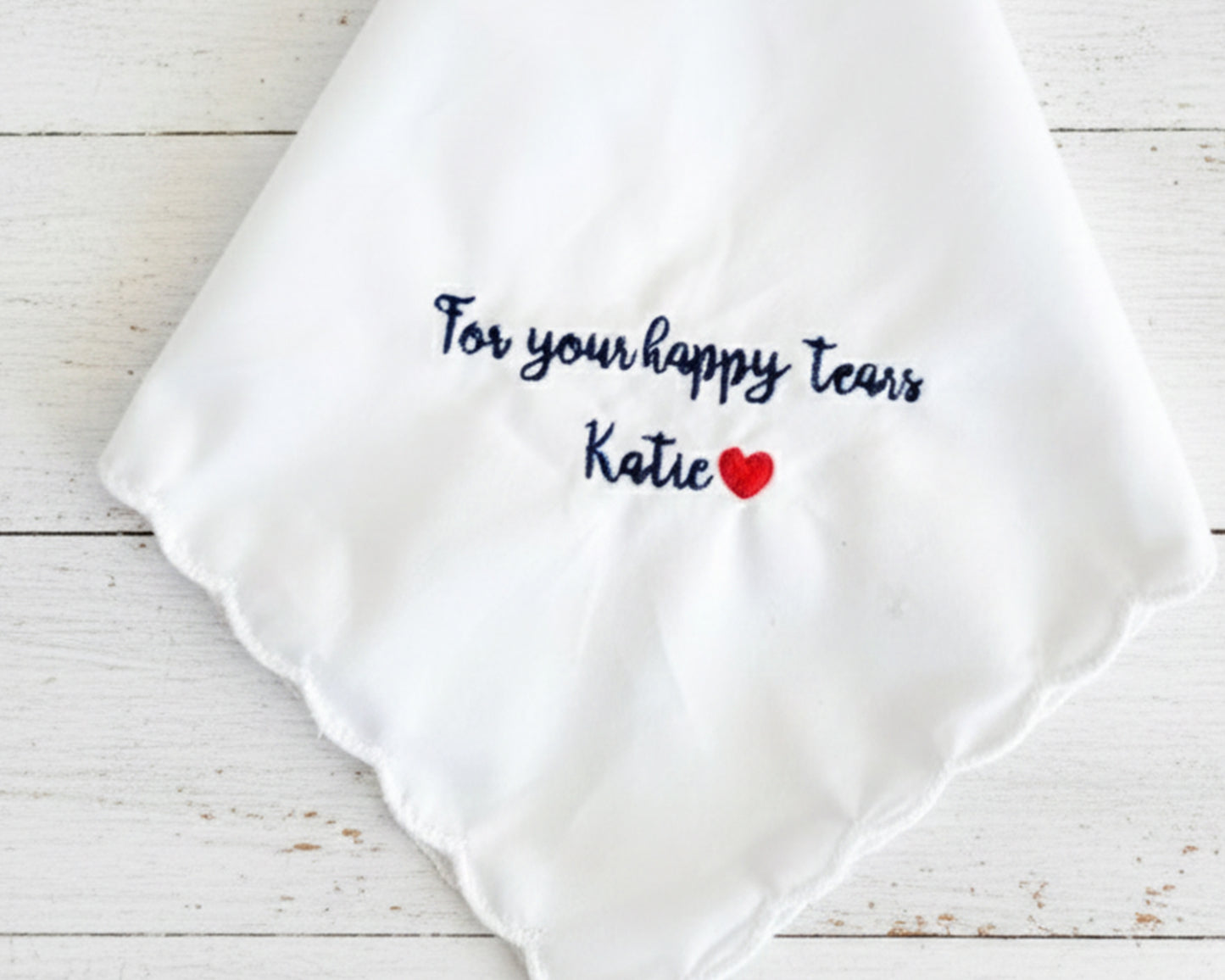 A white scalloped-edge wedding handkerchief made of 100% cotton, featuring navy blue embroidery that reads "For your happy tears Katie" with a small red heart.
