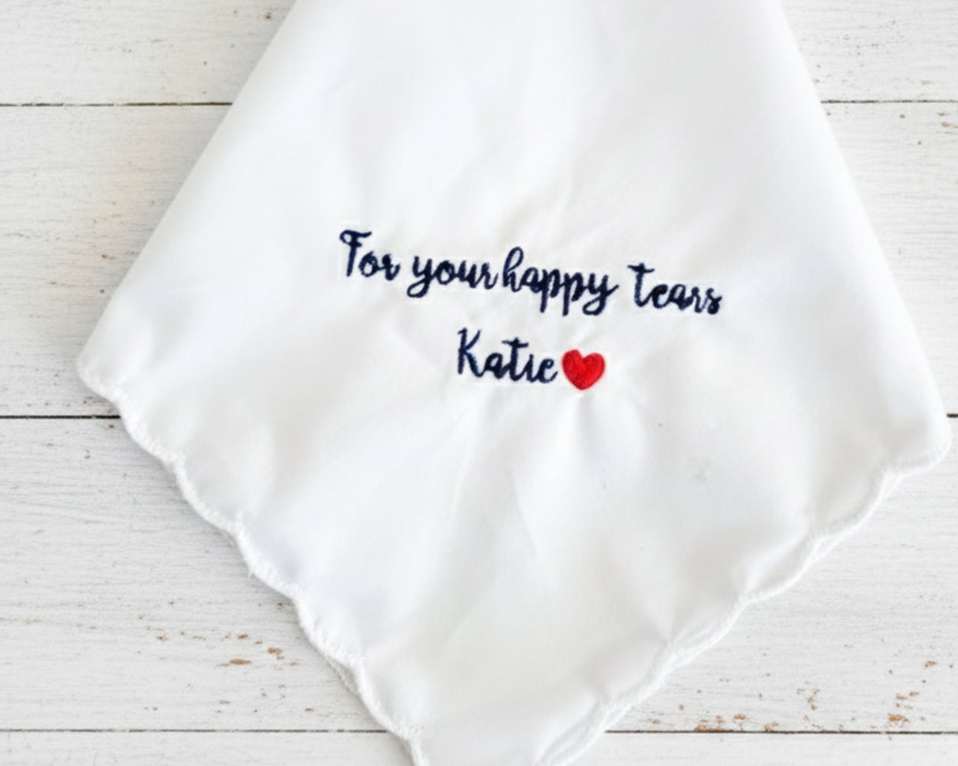 A white scalloped-edge wedding handkerchief made of 100% cotton, featuring navy blue embroidery that reads "For your happy tears Katie" with a small red heart.