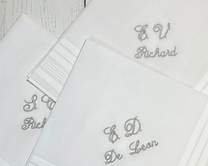 Close-up of three white cotton keepsake handkerchiefs with personalized grey embroidered monograms and script names like "Richard" and "De Leon".