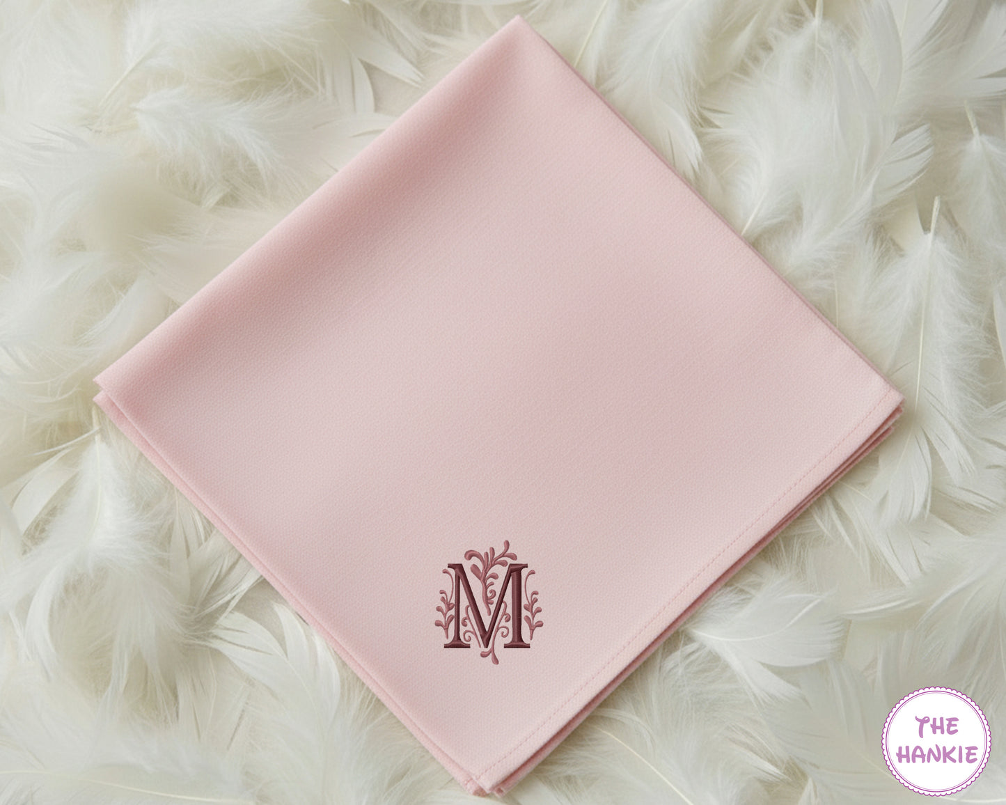A soft pink premium cotton handkerchief featuring a large, decorative embroidered initial "M" with delicate floral accents in a rich plum thread.