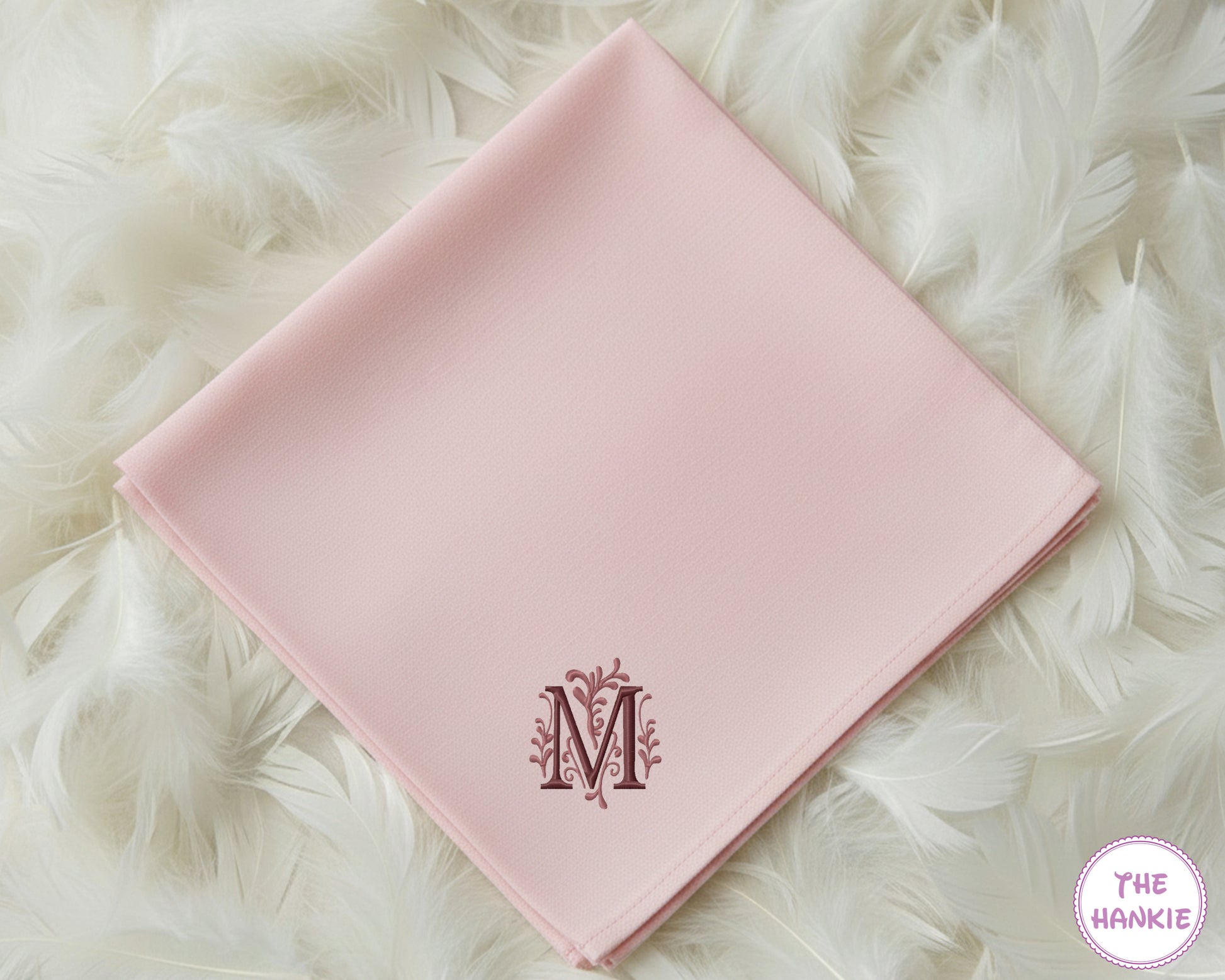 A soft pink premium cotton handkerchief featuring a large, decorative embroidered initial "M" with delicate floral accents in a rich plum thread.