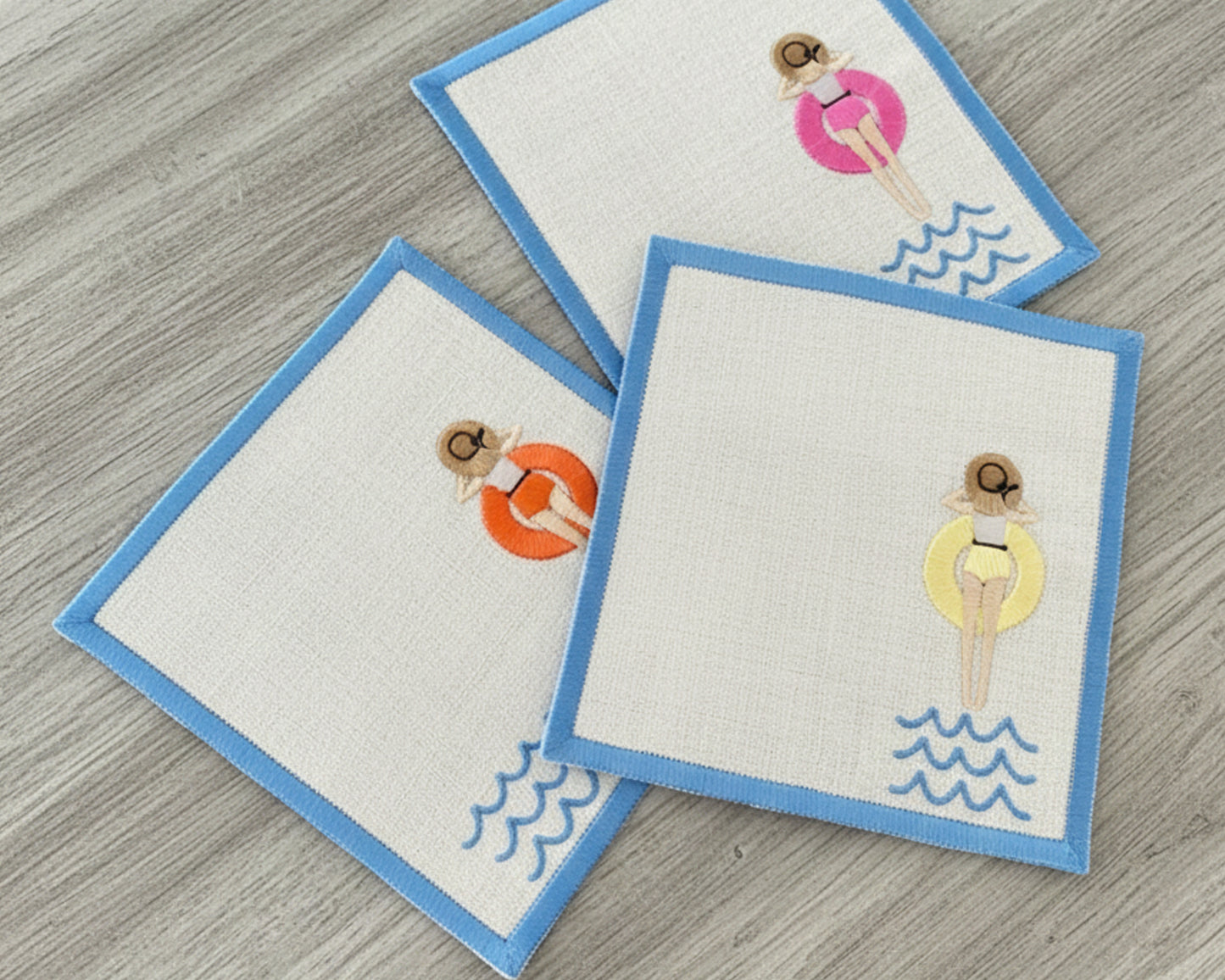 Three white linen napkins on a grey wood surface showing embroidered girls in pink, orange, and yellow pool floats.