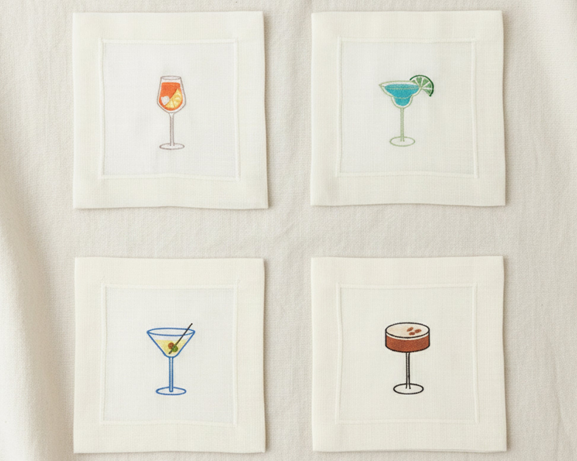 Top-down view of four cotton napkins on a dark marble surface showcasing white wine, Old Fashioned, Gin and Tonic, and Margarita designs.