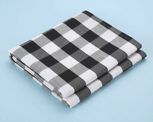 A perfectly folded black and white gingham cotton dinner napkin shown at an angle on a light blue background.