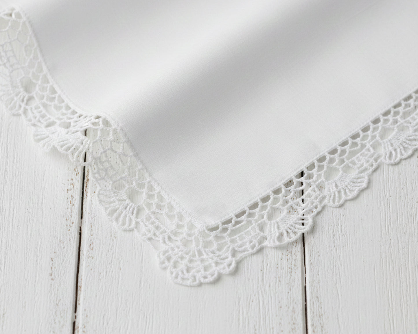Corner detail of a crisp white cotton handkerchief with intricate lace trim on a wood background.