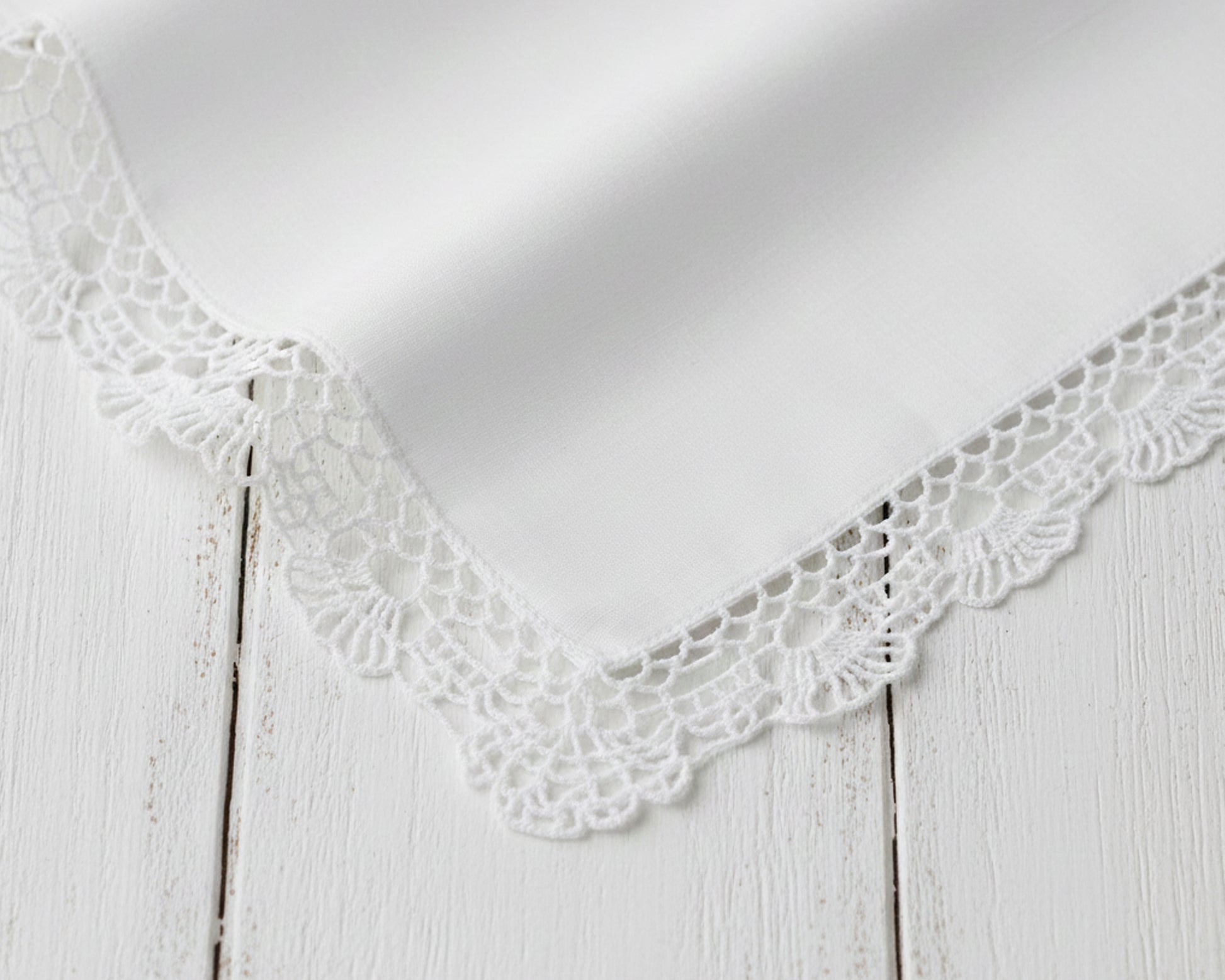 Corner detail of a crisp white cotton handkerchief with intricate lace trim on a wood background.
