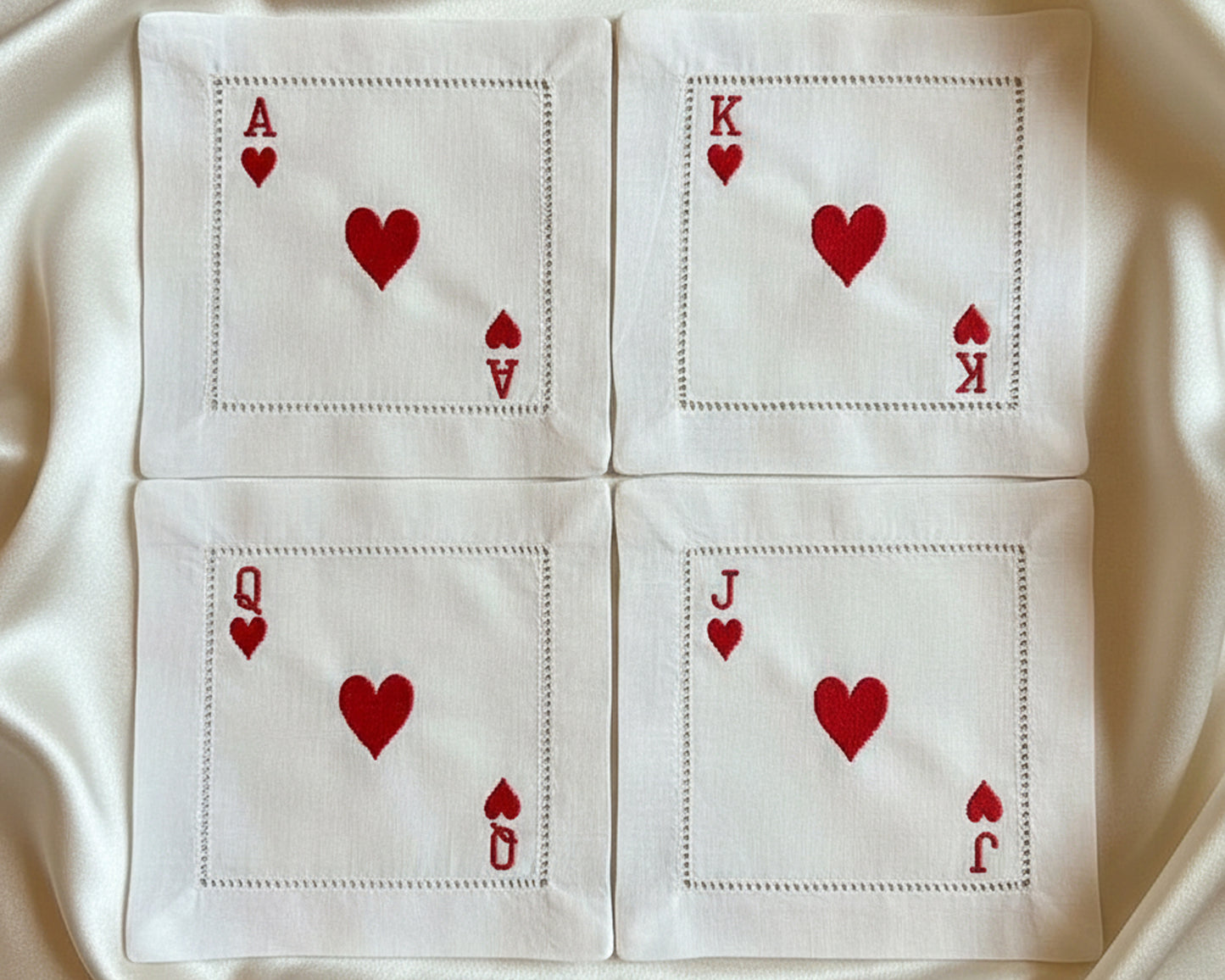 A full set of four white cotton hemstitch napkins featuring the Ace, King, Queen, and Jack of Hearts arranged on a soft silk background.