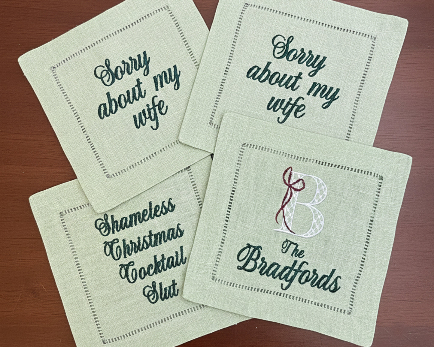 Four sage green linen hemstitch napkins featuring personalized text and monogram embroidery in a dark forest green thread.