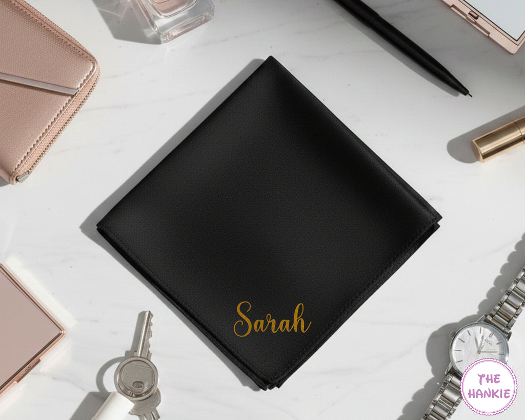 A personalized black cotton pocket square with the name "Sarah" in a vibrant gold glitter vinyl script, shown with luxury accessories.