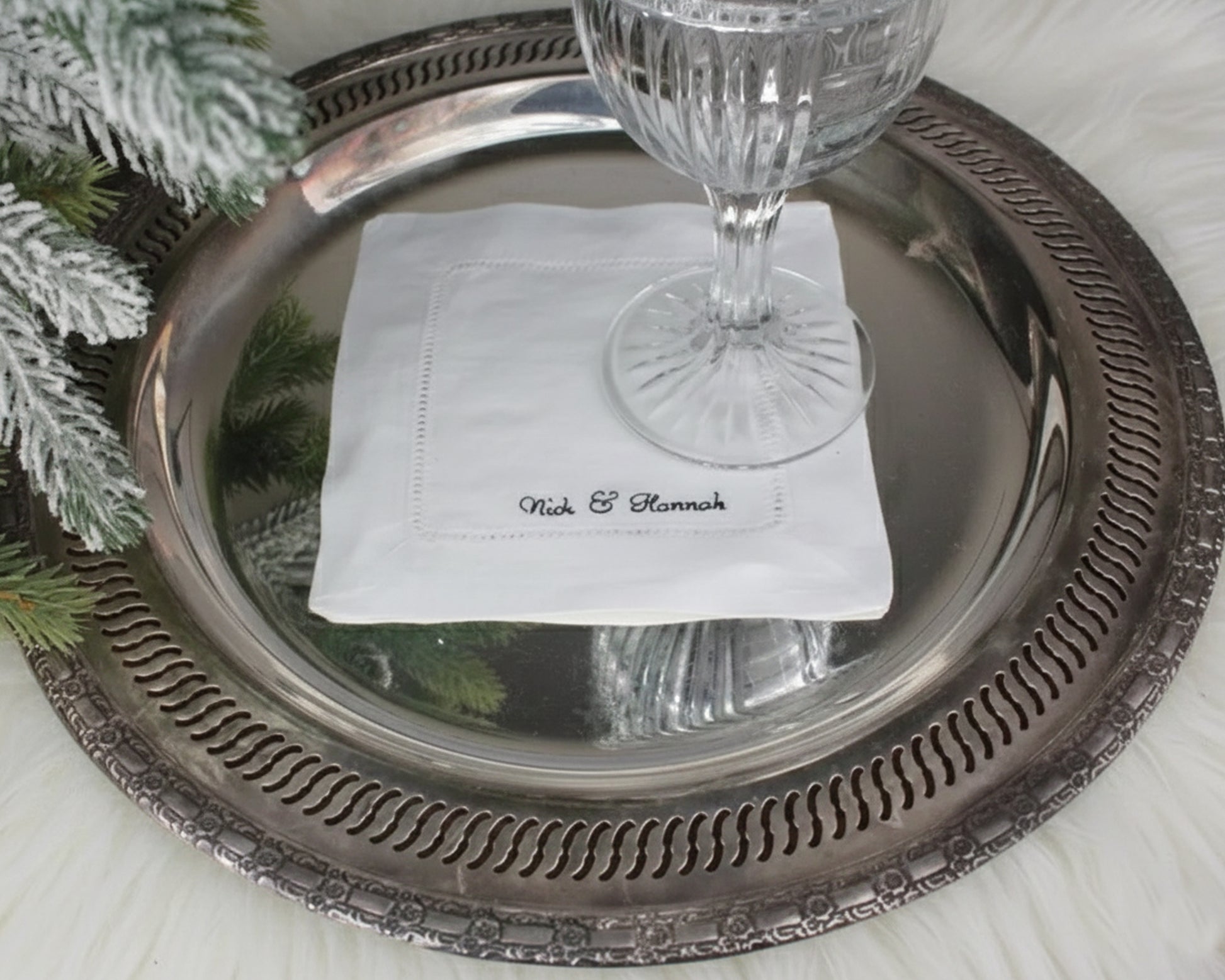 A custom white linen cocktail napkin with a hemstitch border and "Nick & Hannah" embroidery displayed on a vintage silver serving tray.