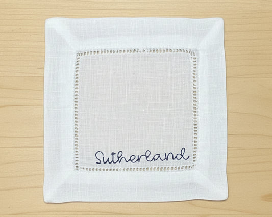 A close-up of a white cotton hemstitch napkin featuring a sophisticated sky blue embroidered letter "B" in a classic serif font.