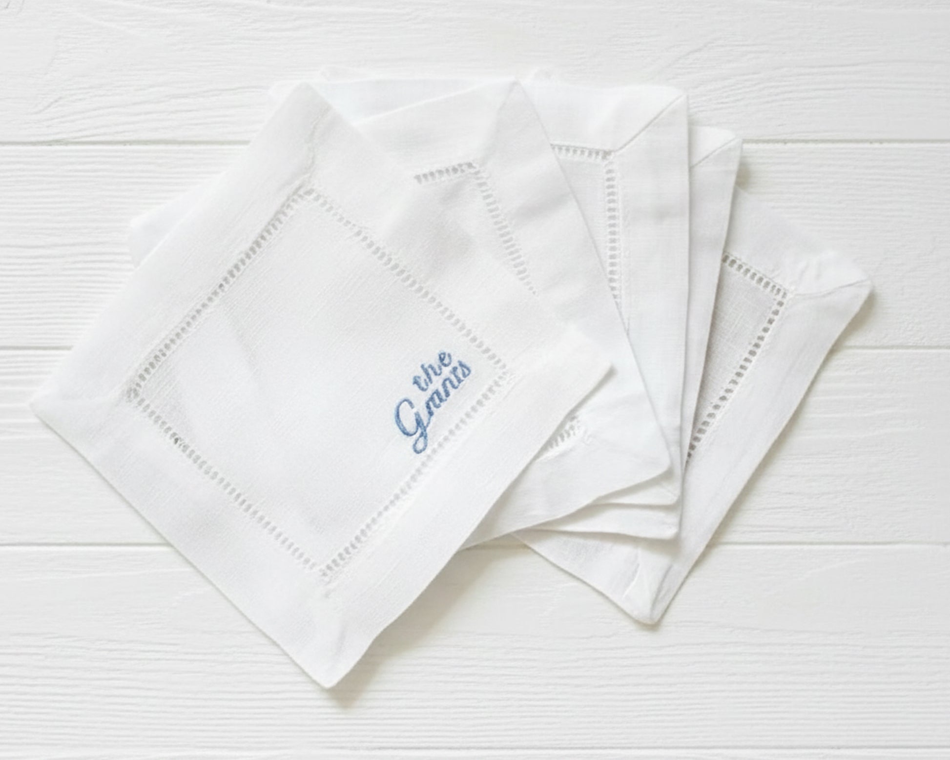 A stack of three premium white cotton cocktail napkins with hemstitch detailing and custom blue script embroidery on a dark marble background.