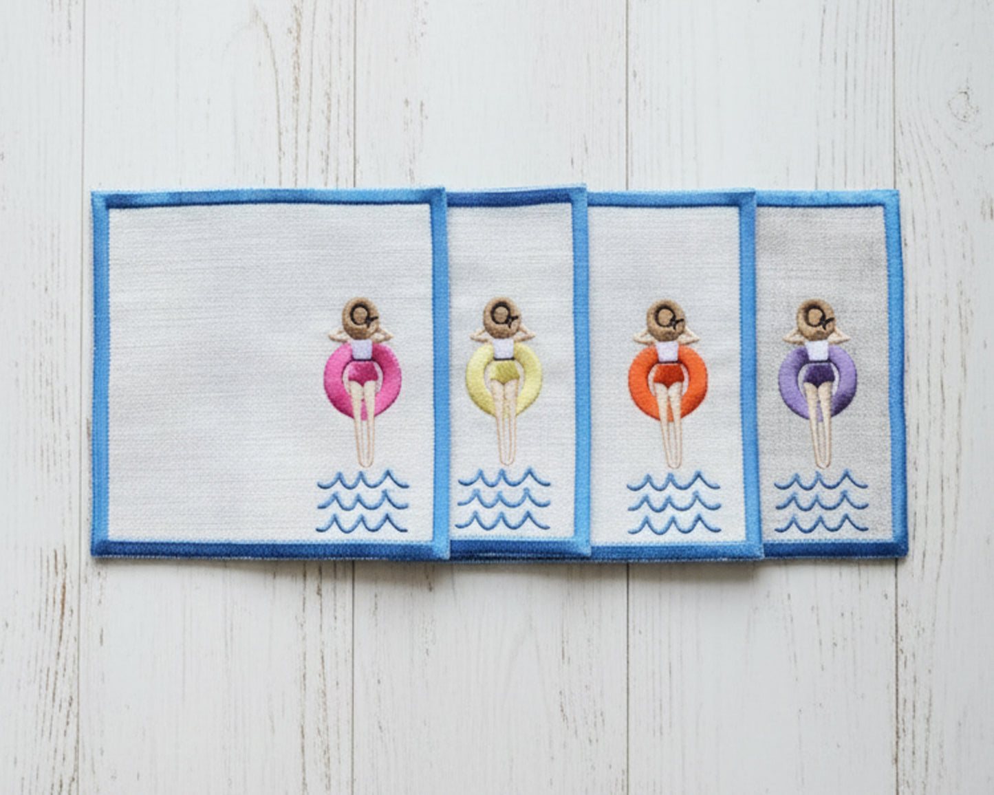 A single sky-blue linen beverage napkin with a scalloped edge and an embroidered girl in a yellow pool floaty on a white fur surface.