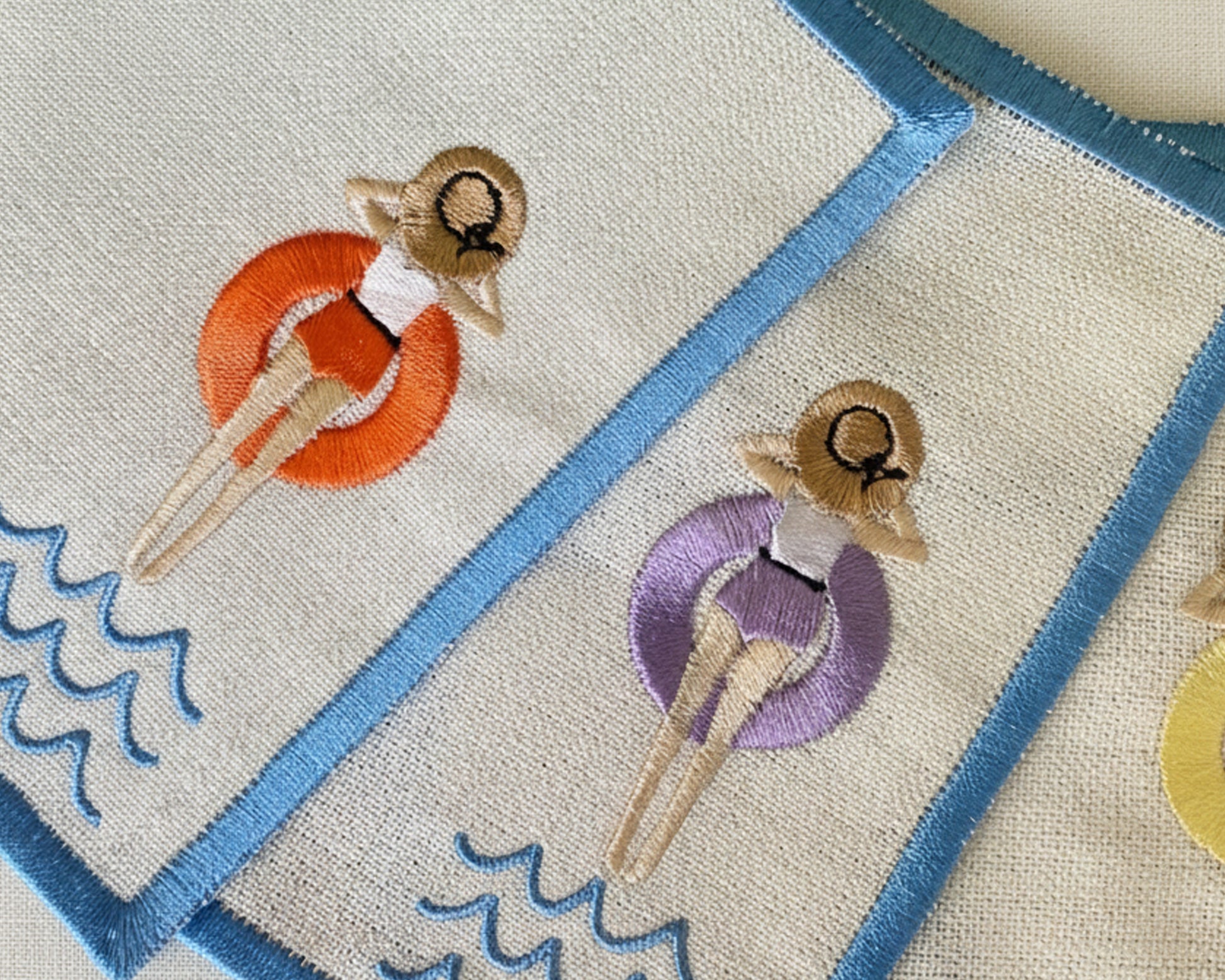 A single sky-blue linen beverage napkin with a scalloped edge and an embroidered girl in a yellow pool floaty on a white fur surface.