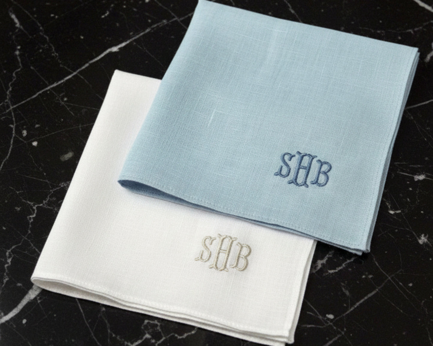 A sky blue and a white linen pocket square featuring custom three-letter monogrammed initials.