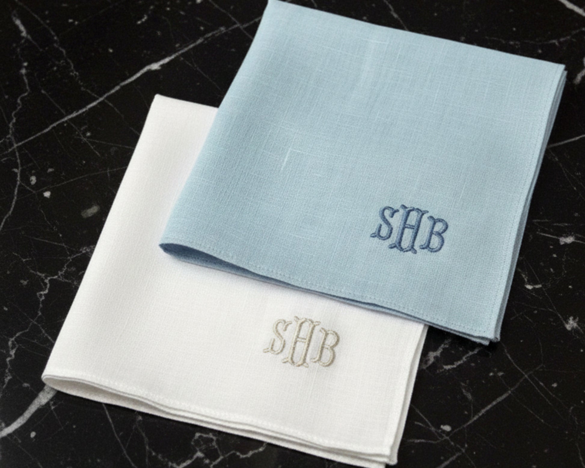 A sky blue and a white linen pocket square featuring custom three-letter monogrammed initials.