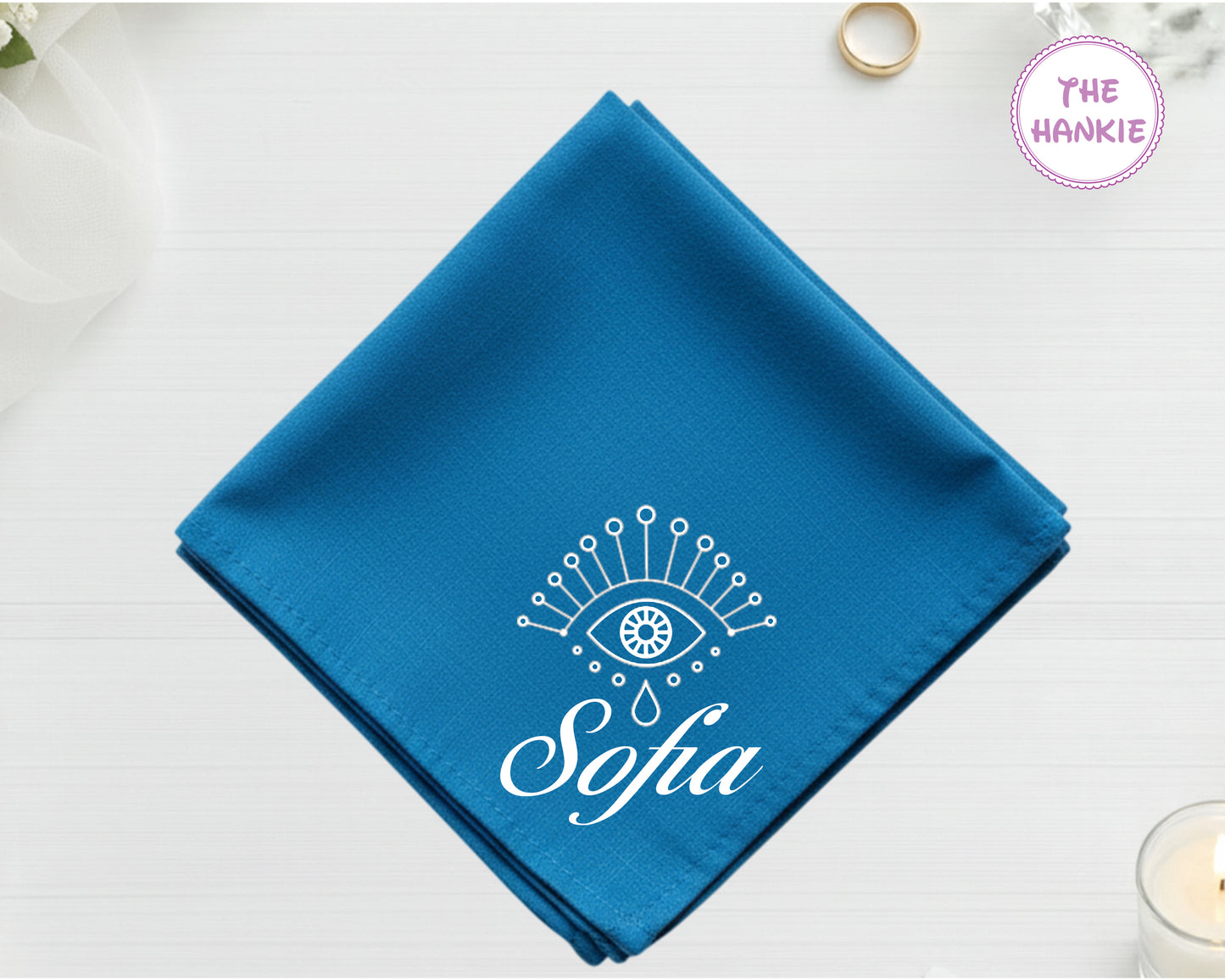 A deep sapphire blue cotton pocket square featuring the name "Sofia" in white script below a white minimalist protective eye with star and droplet details.