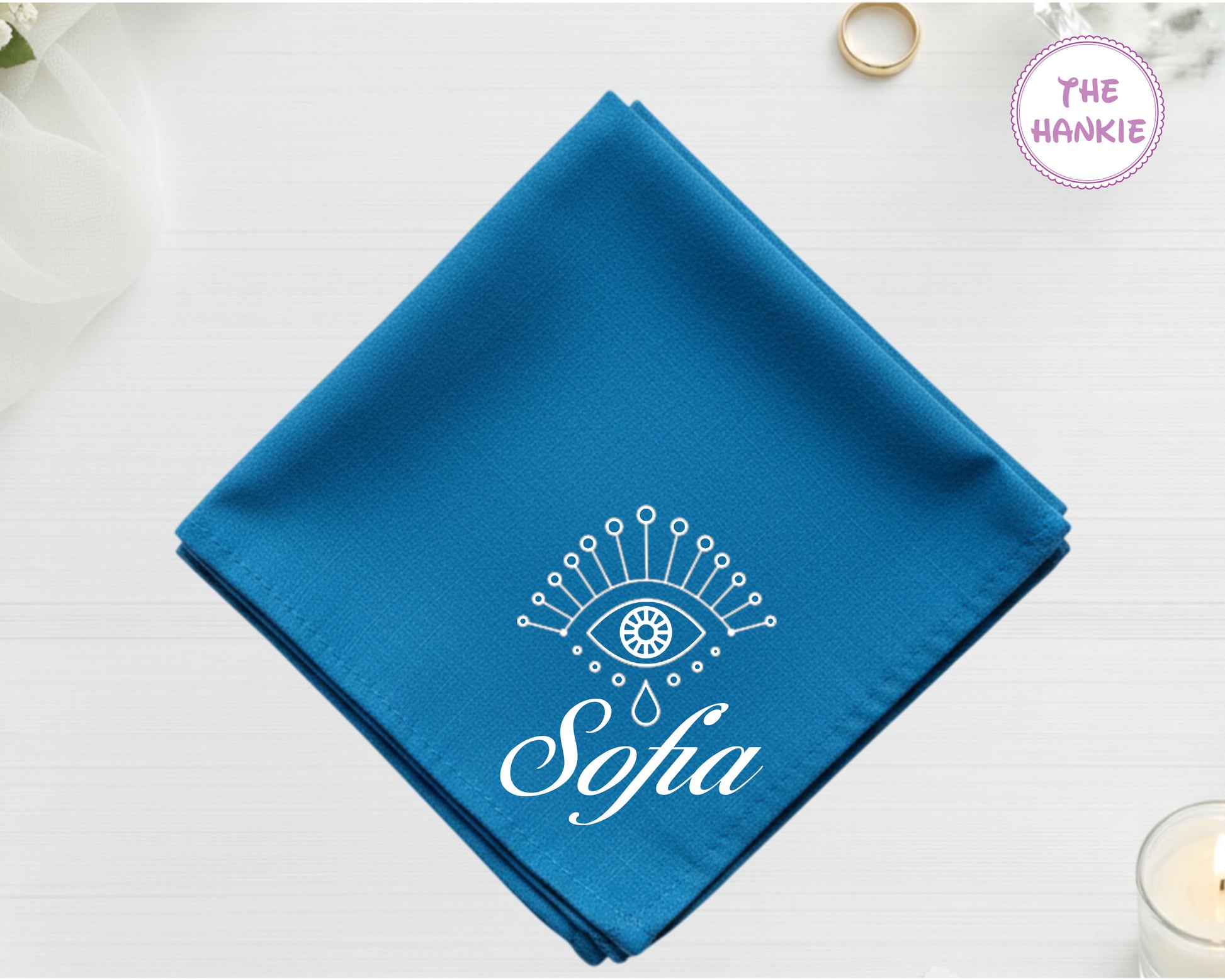 A deep sapphire blue cotton pocket square featuring the name "Sofia" in white script below a white minimalist protective eye with star and droplet details.