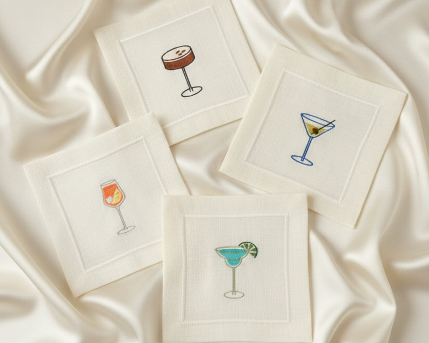 Four 100% cotton napkins displayed on a satin background, showing detailed embroidery of white wine, red wine, a Negroni, and a Margarita.
