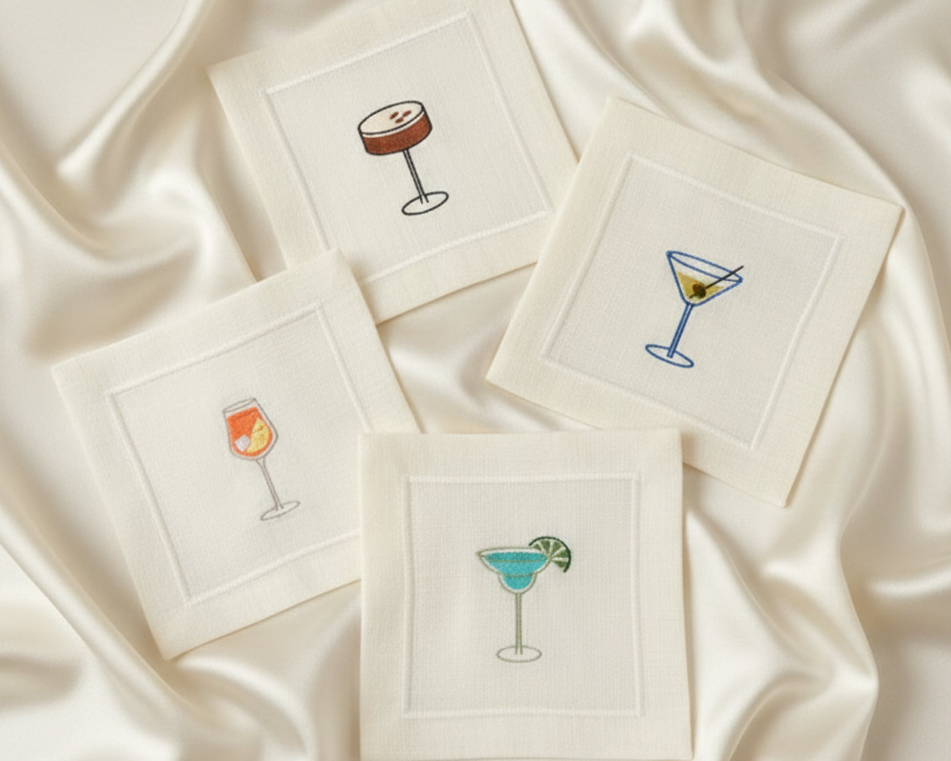 Four 100% cotton napkins displayed on a satin background, showing detailed embroidery of white wine, red wine, a Negroni, and a Margarita.
