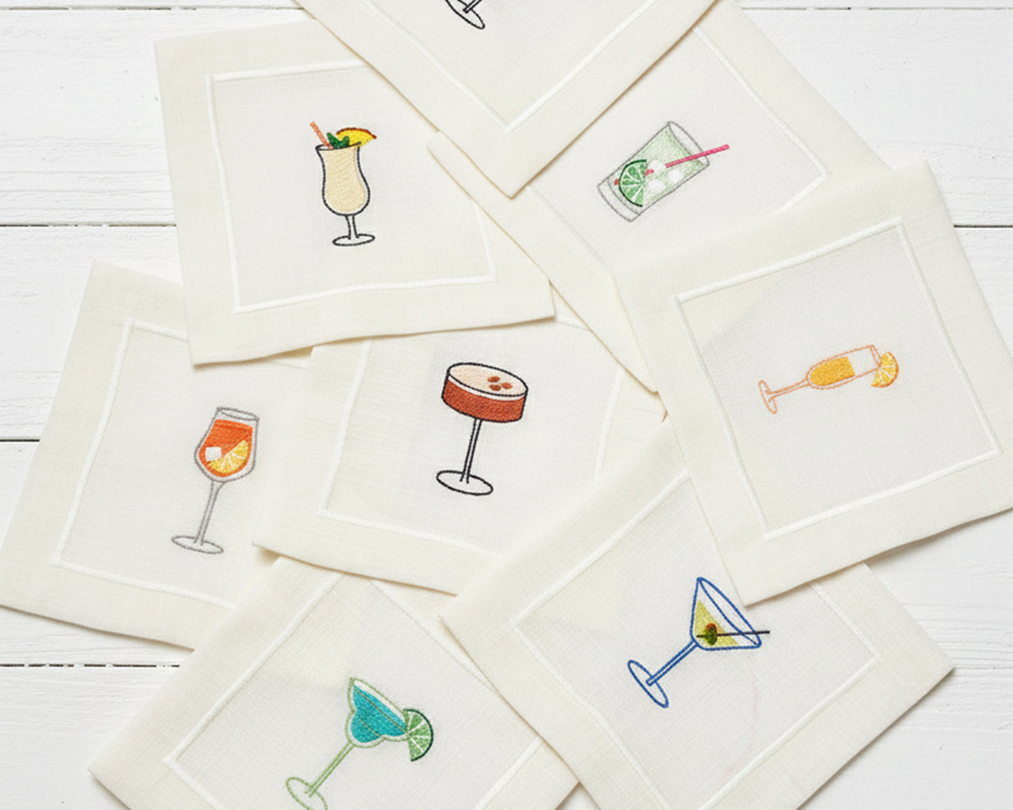 Four 100% cotton napkins displayed on silk, showing detailed embroidery of white wine, red wine, a Negroni, and a Margarita.