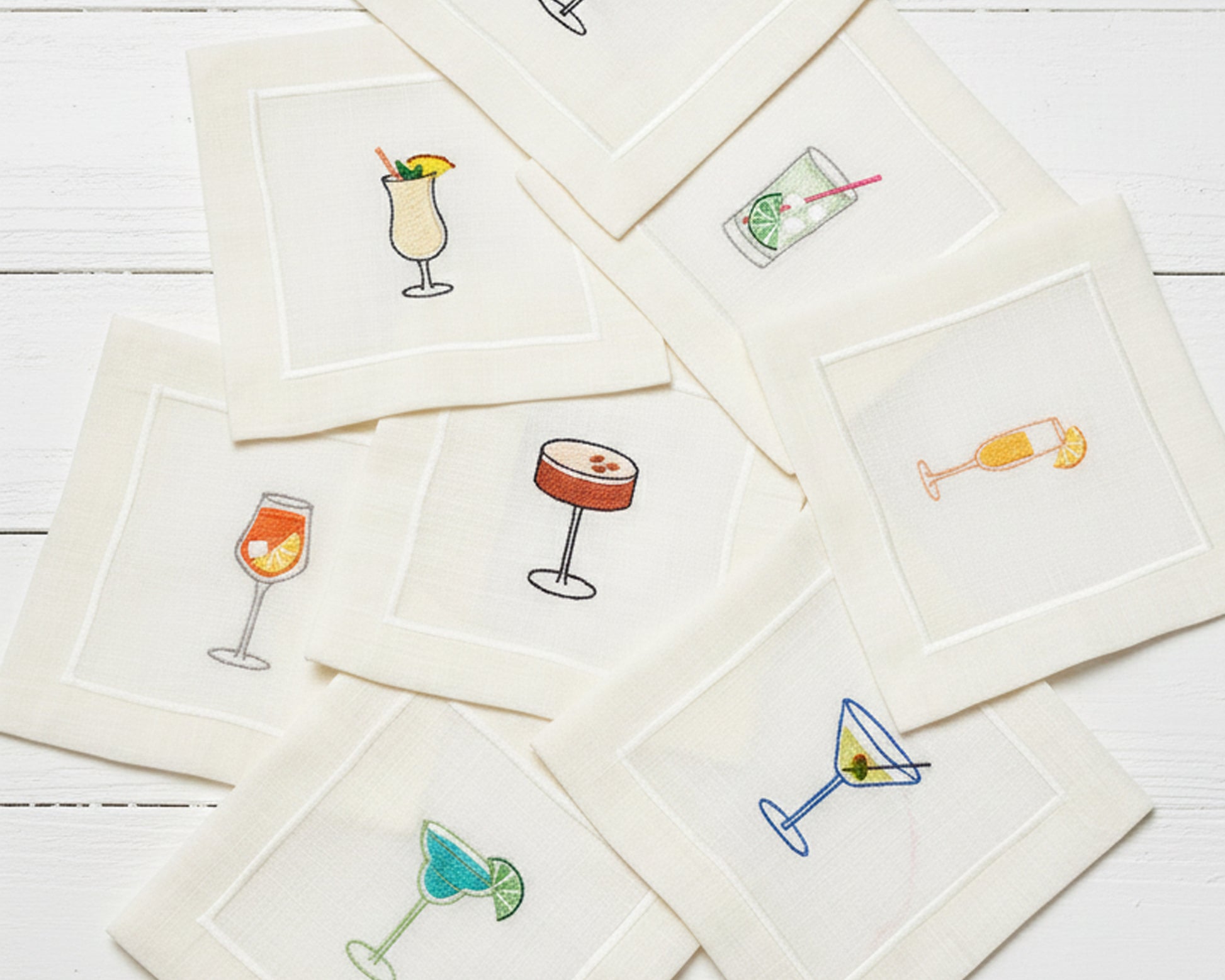 Four 100% cotton napkins displayed on silk, showing detailed embroidery of white wine, red wine, a Negroni, and a Margarita.