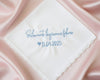 A white scalloped-edge cotton handkerchief with blue embroidery reading "Solamente lágrimas felices 11.04.2023" displayed on elegant cream satin.