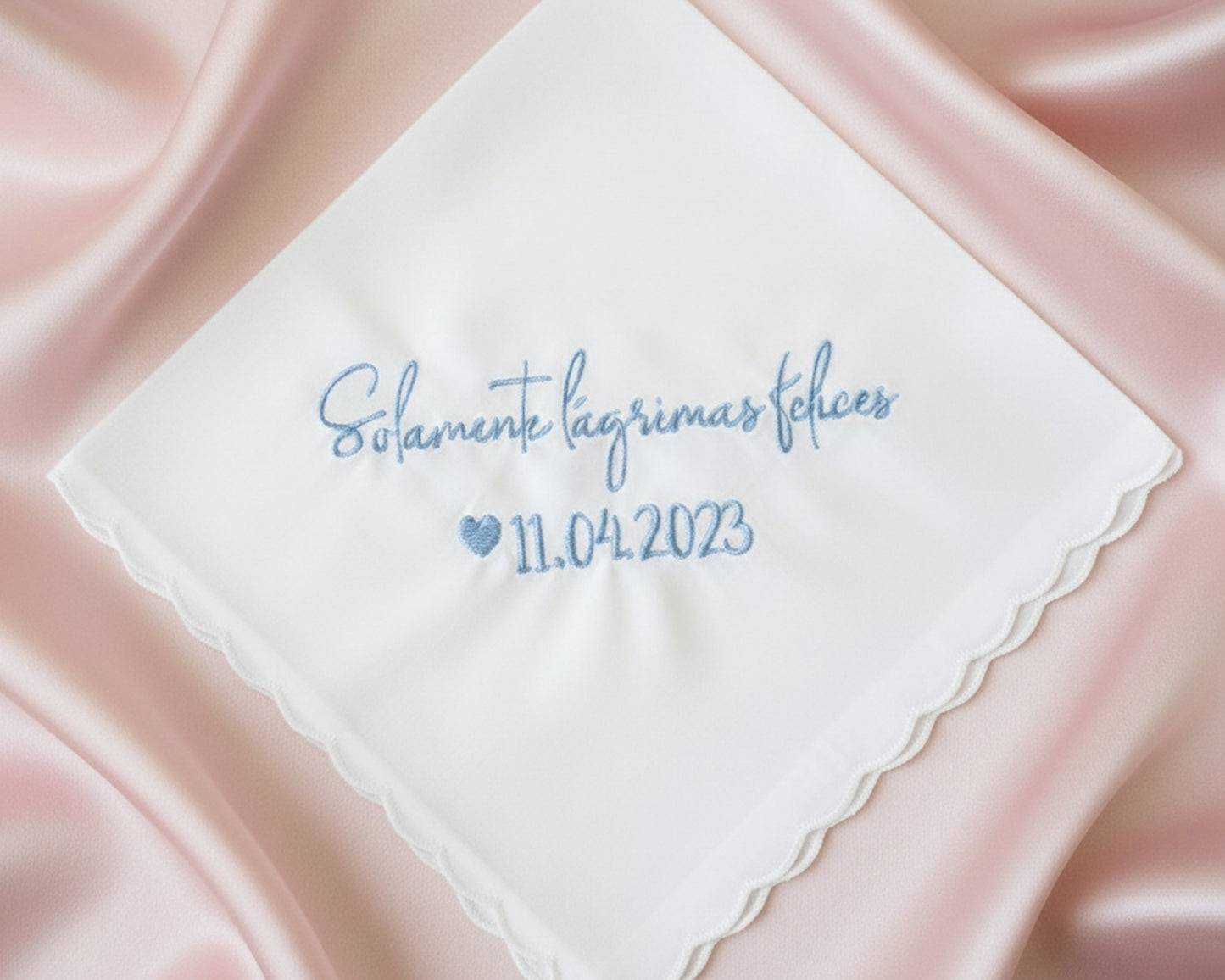 A white scalloped-edge cotton handkerchief with blue embroidery reading "Solamente lágrimas felices 11.04.2023" displayed on elegant cream satin.
