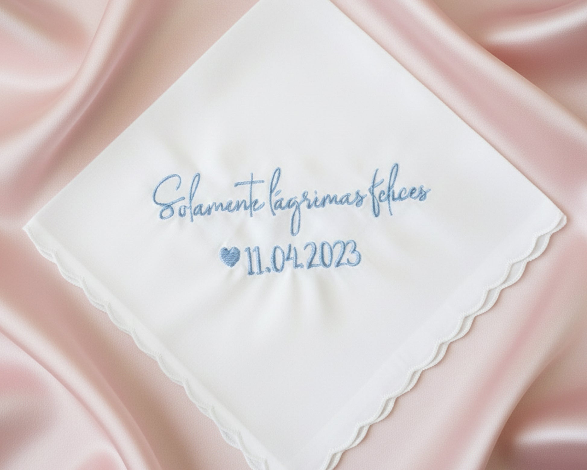 A white scalloped-edge cotton handkerchief with blue embroidery reading "Solamente lágrimas felices 11.04.2023" displayed on elegant cream satin.