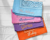 A stack of colorful paisley bandanas custom embroidered with bridesmaid names like Gloria and Abigail on a black marble surface.