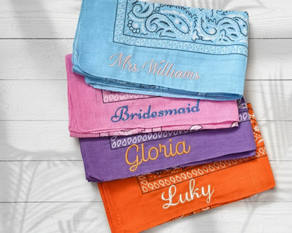 A stack of colorful paisley bandanas custom embroidered with bridesmaid names like Gloria and Abigail on a black marble surface.