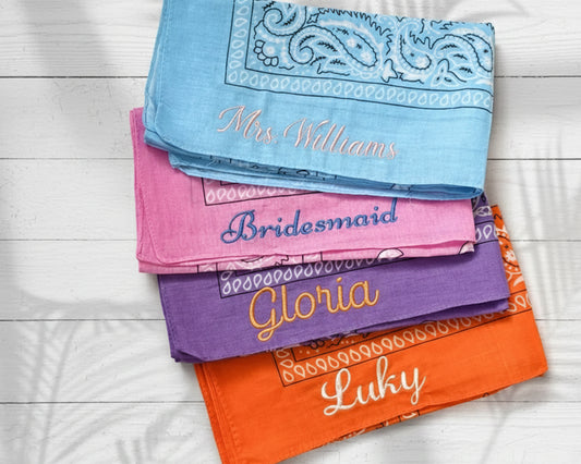 A stack of colorful paisley bandanas custom embroidered with bridesmaid names like Gloria and Abigail on a black marble surface.