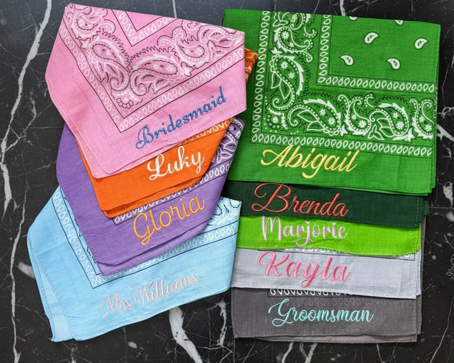 A stack of colorful paisley bandanas custom embroidered with bridesmaid names like Gloria and Abigail on a black marble surface