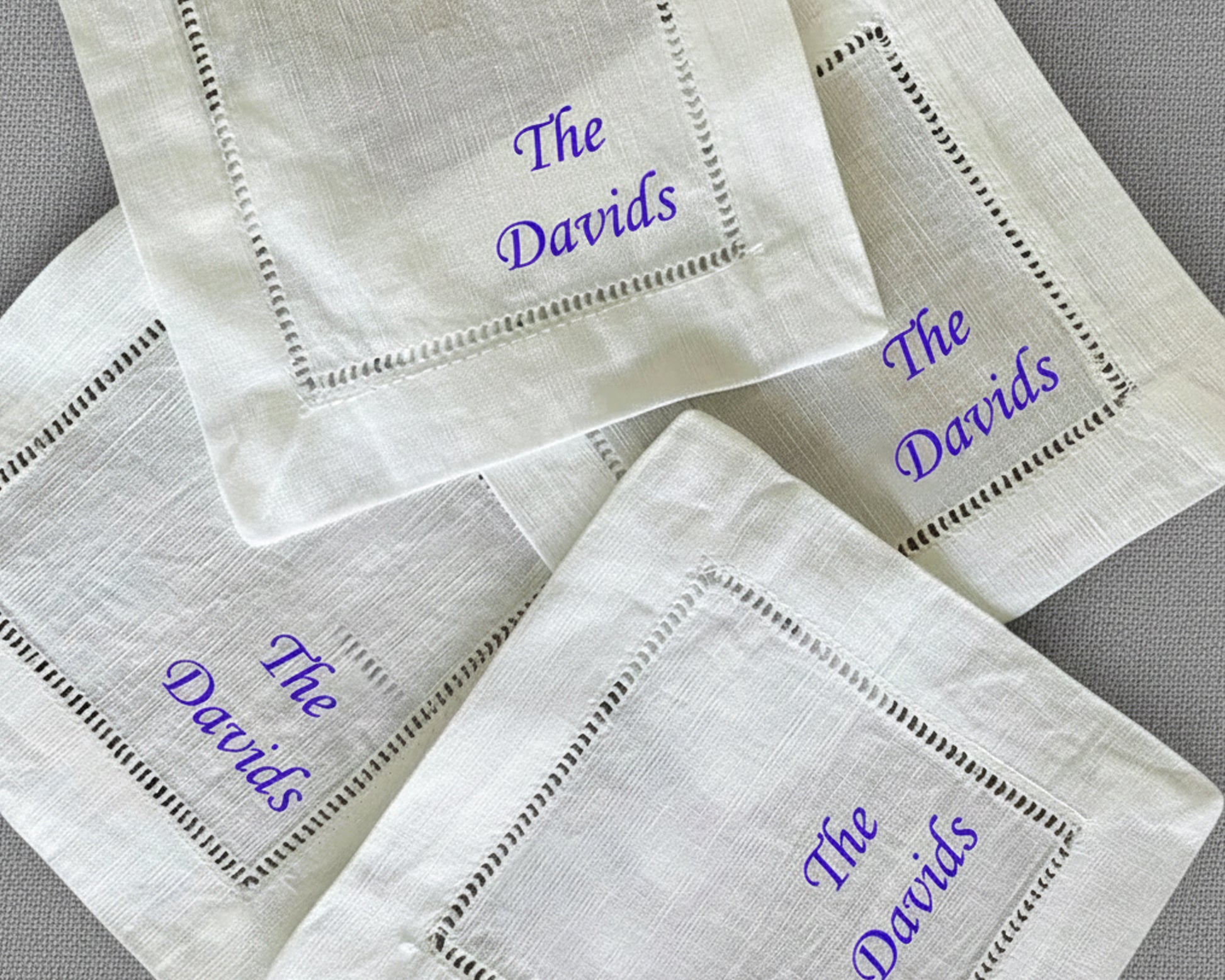 A set of fanned-out white cotton hemstitch napkins featuring vibrant purple script embroidery of the name "The Davids".