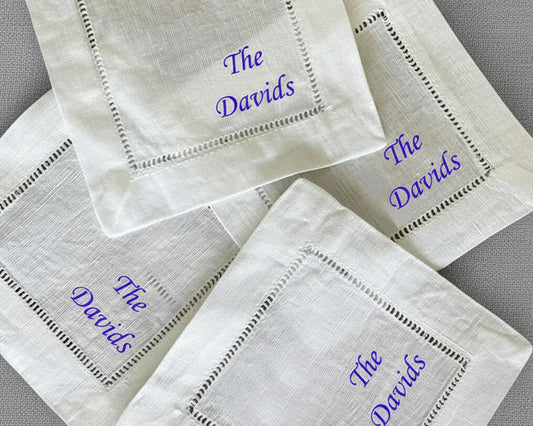 A set of fanned-out white cotton hemstitch napkins featuring vibrant purple script embroidery of the name "The Davids".