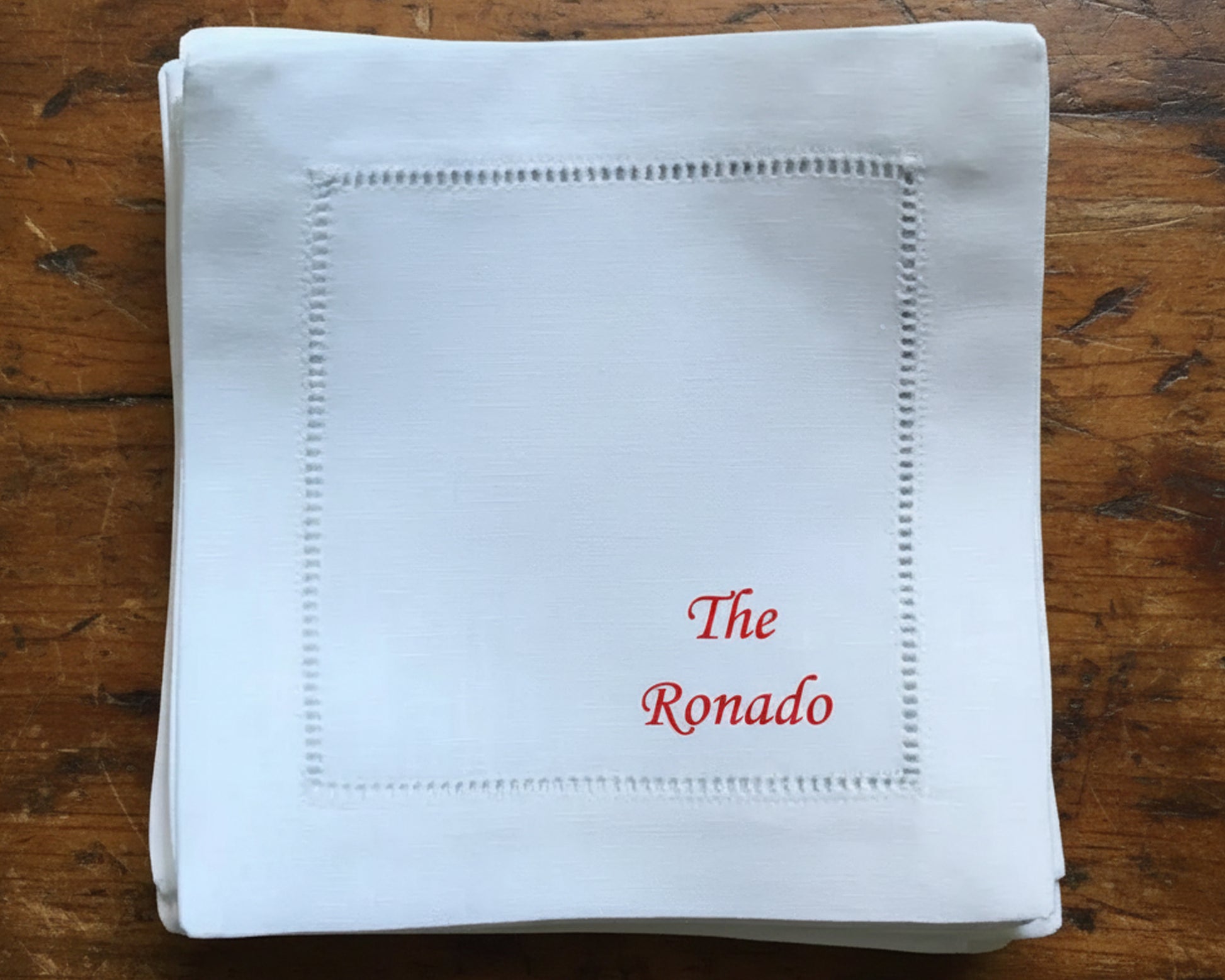 A stack of premium white cotton cocktail napkins with hemstitch detailing and custom red script embroidery for "The Davids" on a black surface.