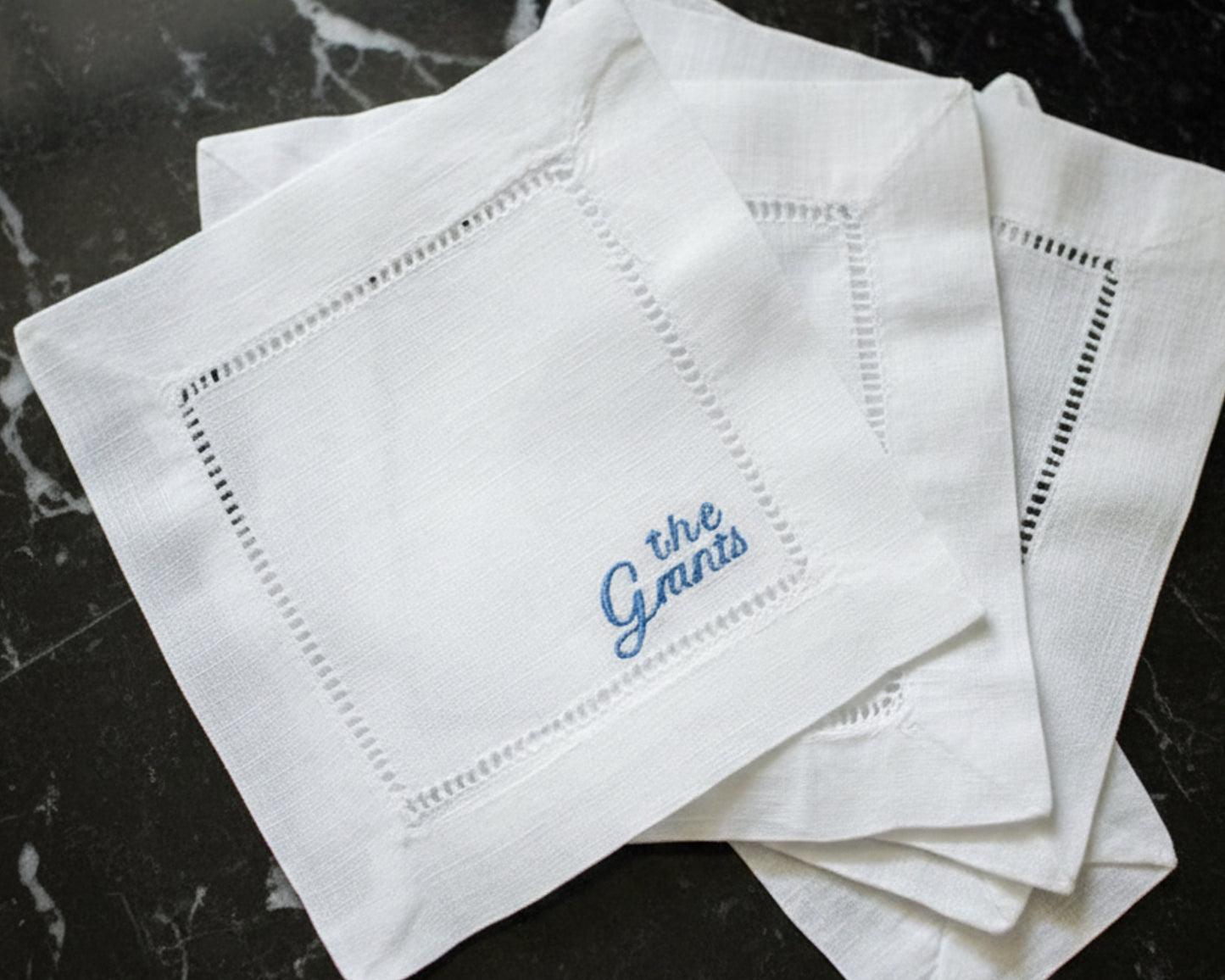 A crisp white 100% cotton hemstitch napkin featuring a sky blue personalized cursive embroidery of the name "the Grants" in a corner layout.