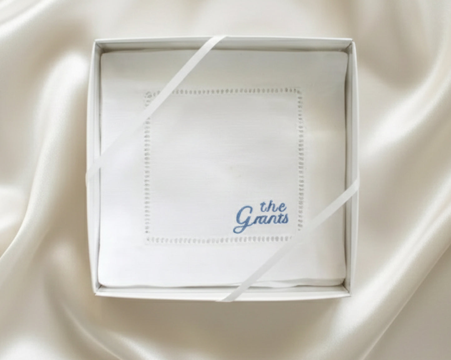 A close-up of a white 100% cotton hemstitch napkin featuring a sky blue personalized heritage script embroidery of the name "the Grants".