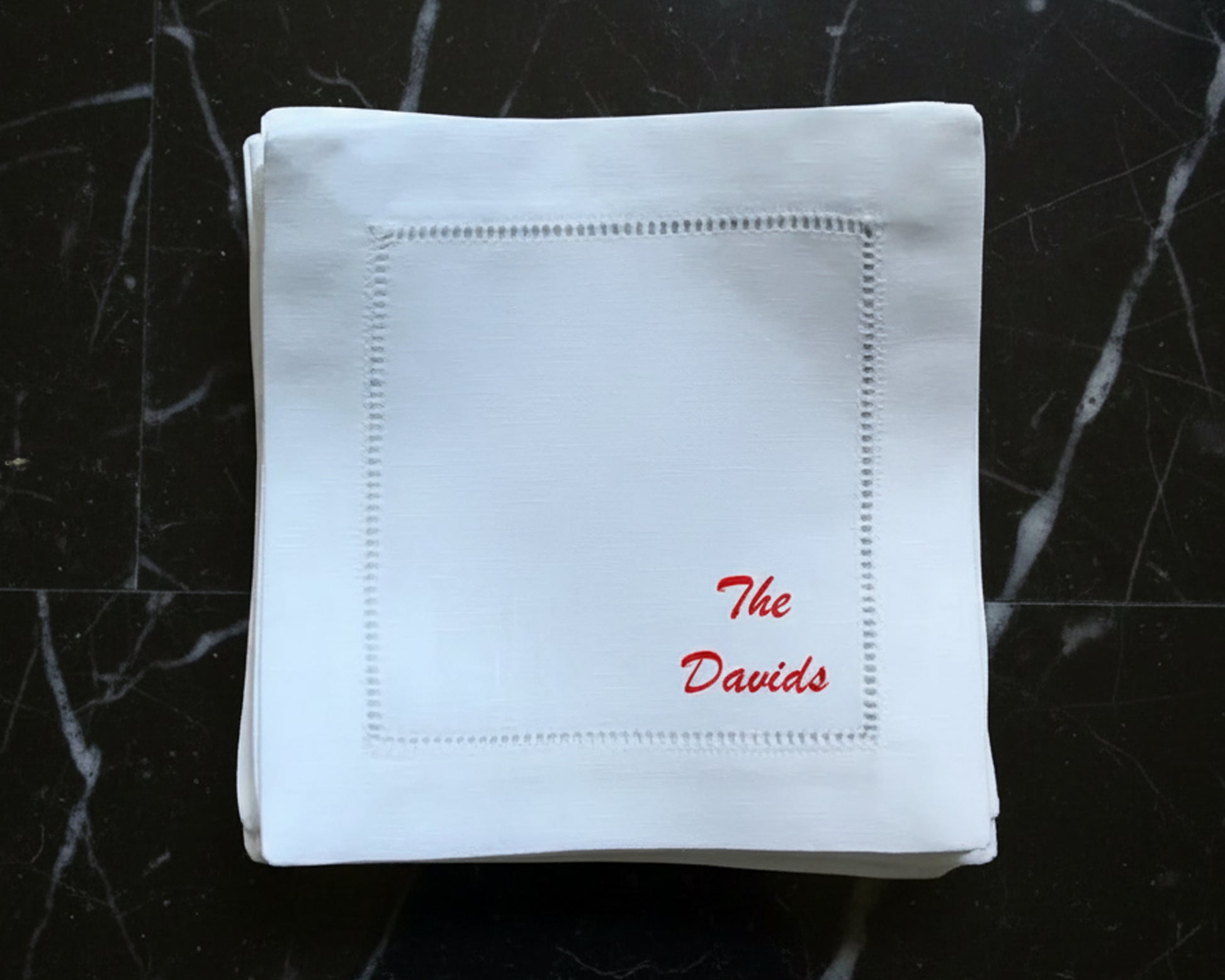 A single white 100% cotton hemstitch napkin featuring a crimson red personalized script embroidery of the name "The Ronado" on a wood background.