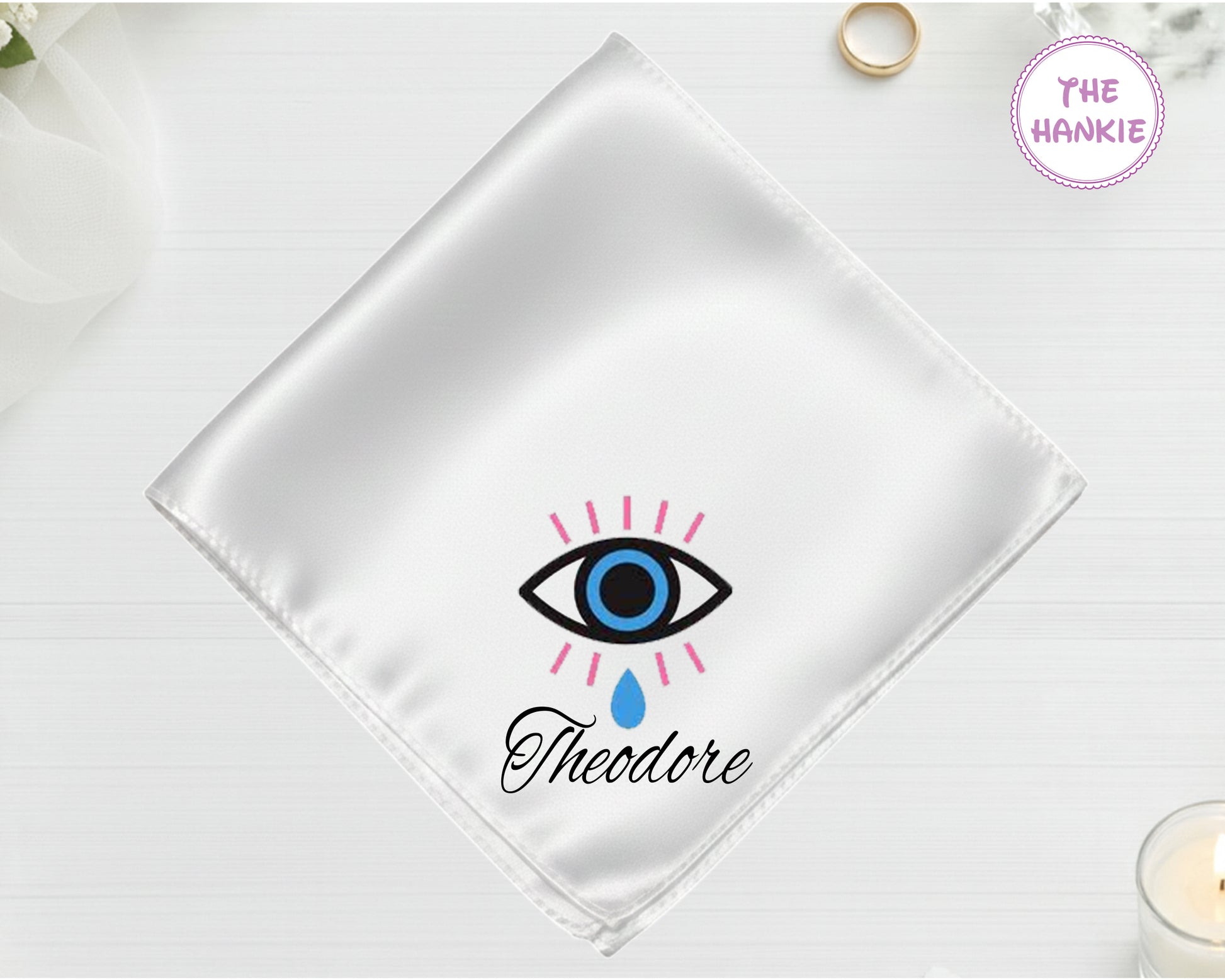 A shimmering pearl white luxury satin handkerchief personalized with "Theodore" below a high-definition vinyl print of a weeping blue evil eye with pink lashes.